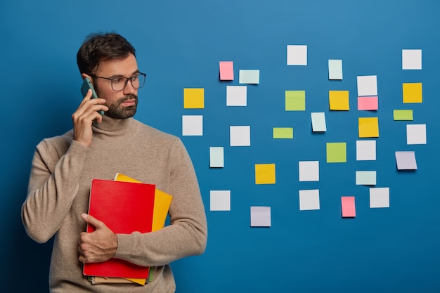 Foto gratuita bel ragazzo barbuto tiene uno smartphone moderno vicino all'orecchio, discute le idee di avvio, vestito con un comodo maglione marrone