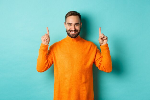 Handsome bearded guy in orange sweater , pointing at top and smiling, standing over turquoise wall.