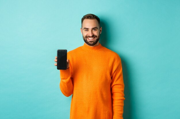 オレンジ色のセーターを着たハンサムなひげを生やした男、スマートフォンの画面と笑顔を表示し、オンラインでプロモーションを表示、ターコイズブルーの壁。