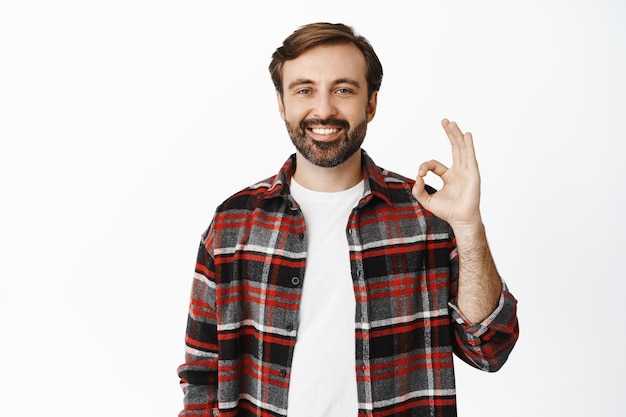 Handsome bearded guy shows okay sign and smiling recommending something good complimenting company standing over white background