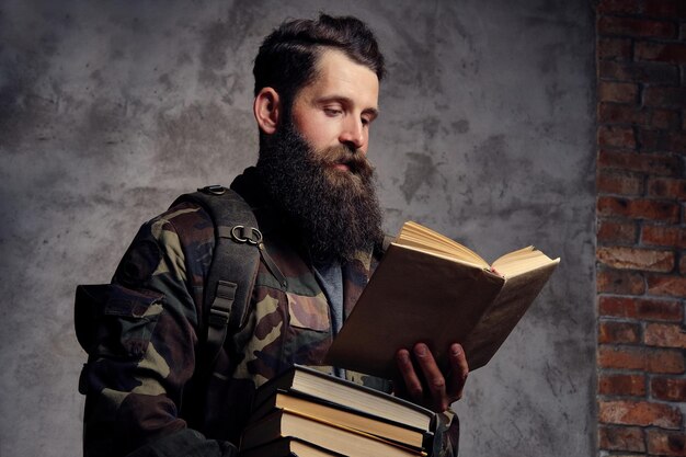 A handsome bearded hipster, dressed in a trendy camouflaged jacket, stands in a studio with a pile of books in his hands and reading, isolated on a dark background.