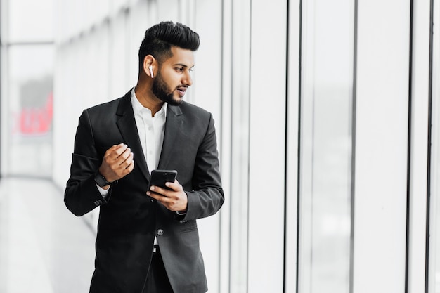 Handsome bearded Indian man, businessman, worker in black suit, he speaks on the phone through wireless headphones