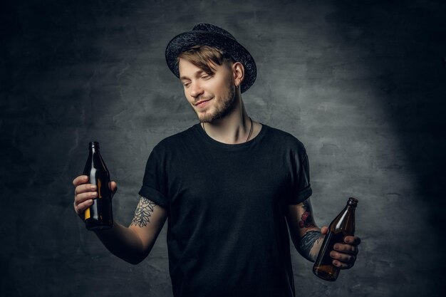 Handsome bearded male with tattooed arms, dressed in a black t shirt and top hat holds craft bottled beer.