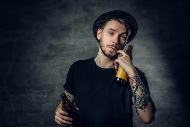 Handsome bearded male with tattooed arms, dressed in a black t shirt and top hat holds craft bottled beer.