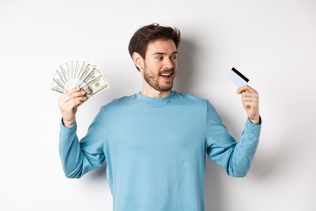 Handsome bearded man choosing between money and plastic credit card, payment with cash or contactless, standing over white background