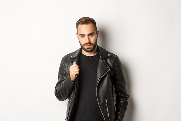 Handsome bearded man expressing confidence, touching his leather jacket and looking self-assured