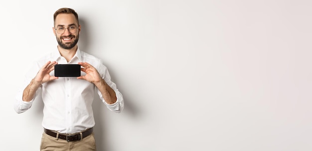 Handsome bearded man in glasses demonstrating mobile phone application showing smartphone screen sta