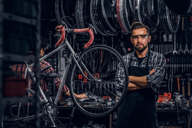 眼鏡をかけたハンサムなひげを生やした男が、自分のワークショップで固定自転車の近くに立っています。