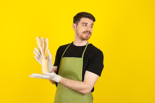 Handsome bearded man holding wooden spoons and looking away on a yellow.