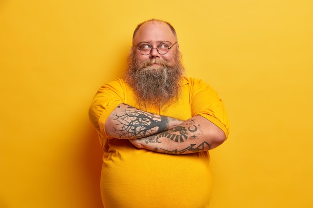 Free photo handsome bearded man keeps arms folded, looks thoughtfully away, has plump body, dressed in casual clothes, makes up plan how to lose weight, isolated over yellow wall. pensive indecisive guy