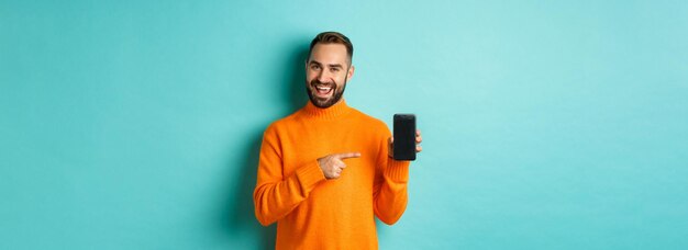 Красивый бородатый мужчина в оранжевом свитере, указывающий пальцем на экран мобильного телефона, показывающий приложение sm