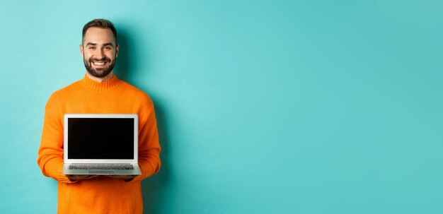 Красивый бородатый мужчина в оранжевом свитере, показывающий экран ноутбука, демонстрирующий интернет-магазин, стоящий на улице