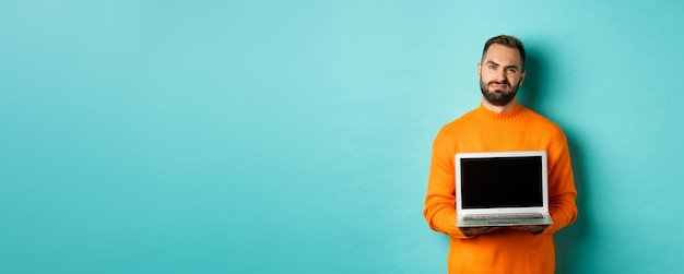 Free photo handsome bearded man in orange sweater showing laptop screen demonstrating promo grimacing disappoin