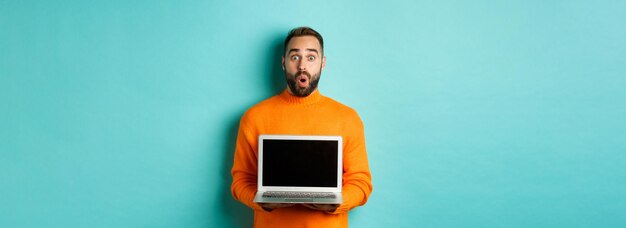 빛 위에 서있는 프로모션을 보여주는 노트북 화면을 보여주는 주황색 스웨터를 입은 잘 생긴 수염 난 남자