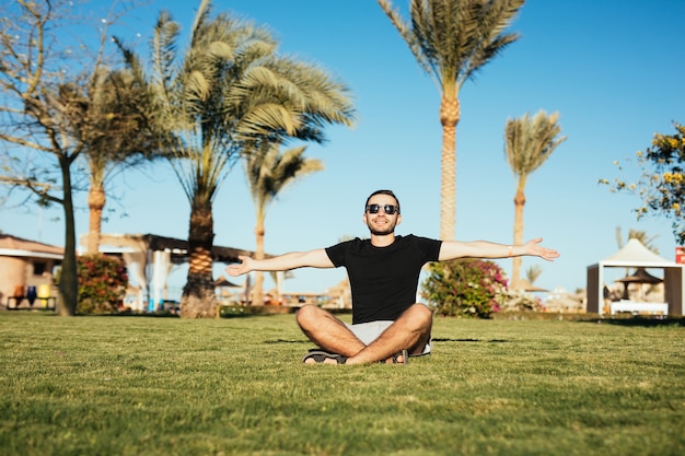 Handsome bearded man in sunglasses sitting on green grass and rise palms to sun.
