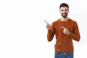 Free photo handsome bearded man in sweatshirt, pointing fingers left at copy space blank smiling pleased, advice buy subscription, click link or follow page to find out info, white wall