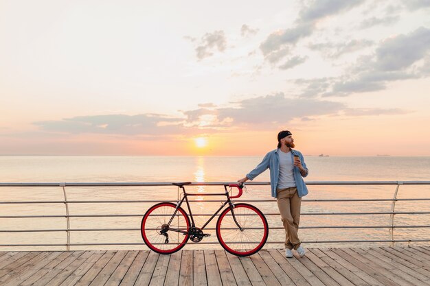 Handsome bearded man traveling with bicycle in morning sunrise by the sea drinking coffee, healthy active lifestyle traveler