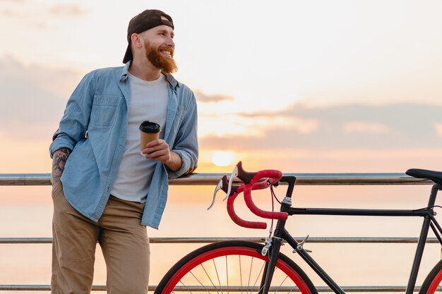 Handsome bearded man traveling with bicycle in morning sunrise by the sea drinking coffee, healthy active lifestyle traveler