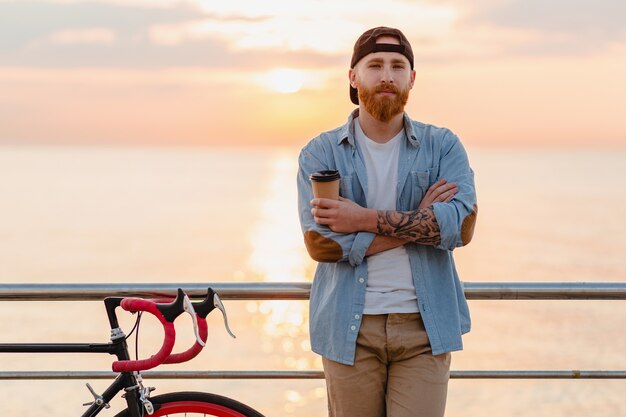 Красивый бородатый мужчина, путешествующий с велосипедом в утреннем восходе солнца у моря, пьет кофе, путешественник здорового активного образа жизни