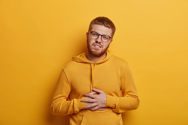 Handsome bearded man with ginger hair feels stomachache, suffers from indigestion and abdomainal cramps after eating spoiled product, wears glasses and sweatshirt, isolated on yellow wall