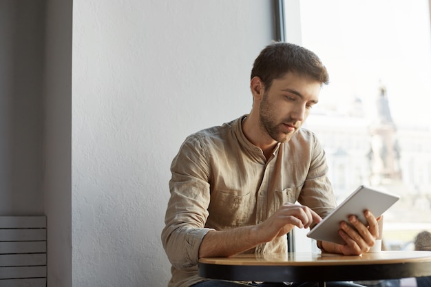 Handsome bearded man with short hair in casual clothes sitting in cafe, looking through startup project details on tablet. Business concept.