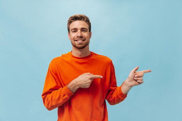 Handsome bearded man with stylish hairstyle in bright orange sweatshirt looking into camera smiling and pointing to place for text