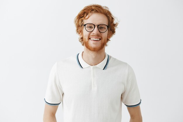 Handsome bearded redhead guy posing against the white wall with glasses