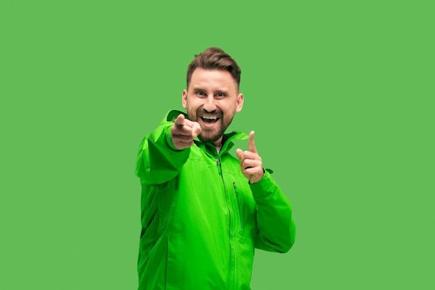 Handsome bearded smiling happy young man looking and pointing at camera isolated on vivid trendy green studio. concept of the autumn and cold time. Human emotions concepts