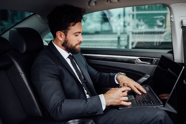Красивый, бородатый, улыбающийся топ-менеджер в черном костюме работает на своем ноутбуке на заднем сиденье автомобиля