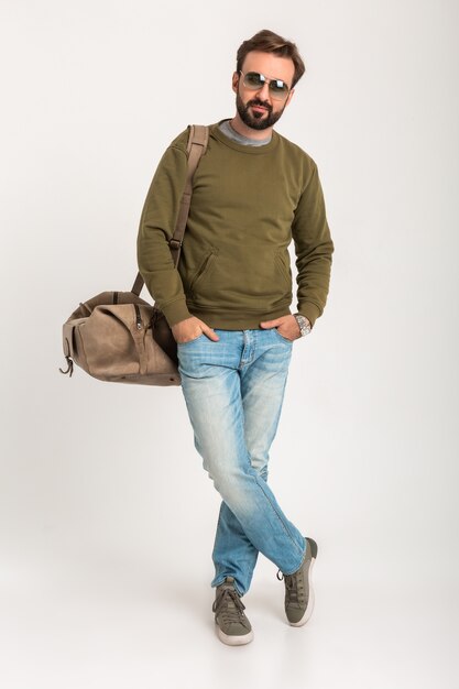 Красивый бородатый стильный мужчина позирует изолированно, одетый в толстовку с дорожной сумкой, в джинсах и солнцезащитных очках