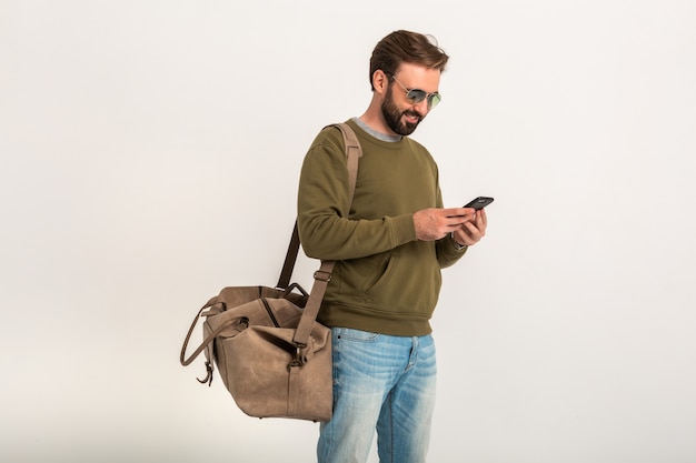 Handsome bearded stylish man in sweatshirt with travel bag, wearing jeans and sunglasses isolated holding phone