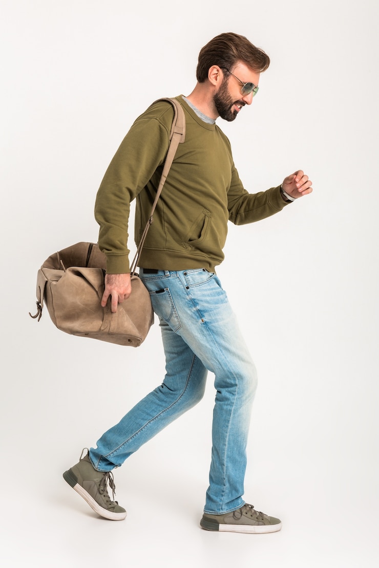 Free Photo handsome bearded stylish man walking isolated dressed in sweatshirt with travel bag, wearing jeans and sunglasses