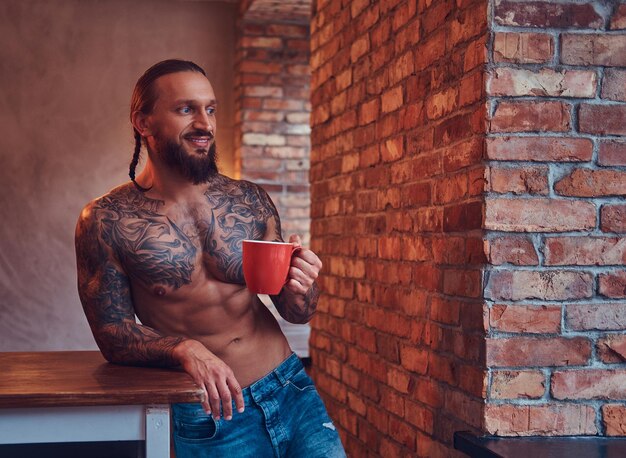 Handsome bearded tattoed male with a stylish haircut and muscular body, drinks coffee, leaning on a table.