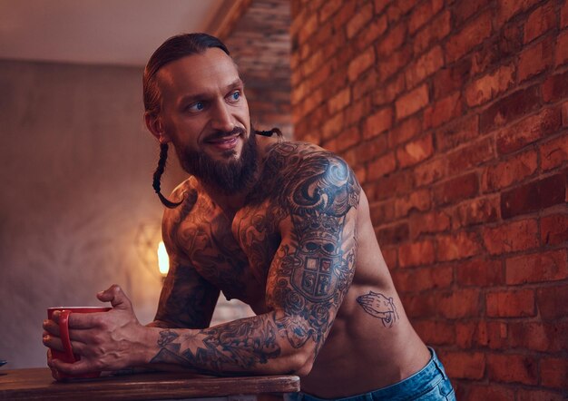 Handsome bearded tattoed male with a stylish haircut and muscular body, drinks coffee, leaning on a table.