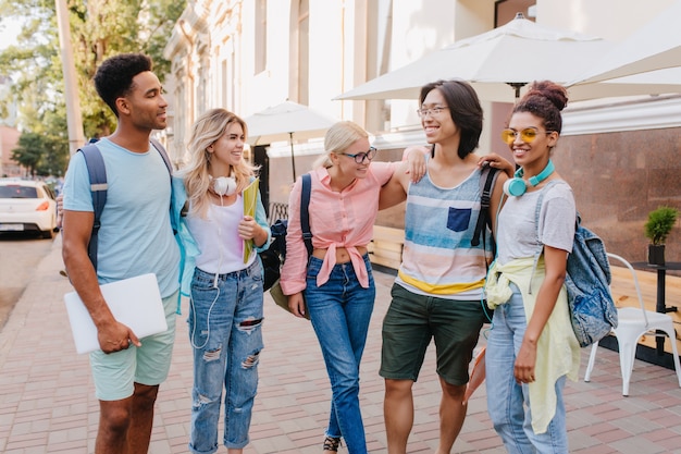 Handsome black guy with laptop looking at asian boy in glasses, which gently embracing cute girls. Students in stylish clothes hanging out together on the street and laughing.