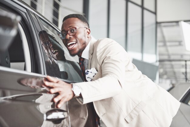 ディーラーでハンサムな黒人男性が彼の新しい車を抱いて、笑っています。