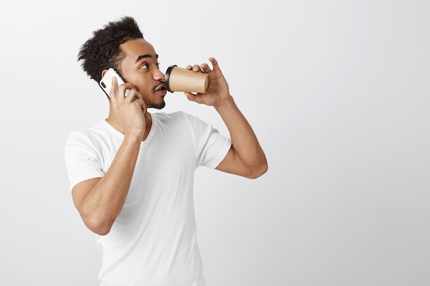 Красивый темнокожий мужчина в белой футболке разговаривает по мобильному телефону и пьет кофе на вынос, глядя вправо