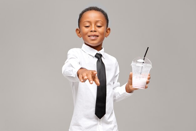 красивый черный школьник в рубашке и галстуке держит прозрачное пластиковое стекло, пьет здоровый энергетический протеиновый молочный коктейль с счастливым довольным выражением лица. Здоровье и еда