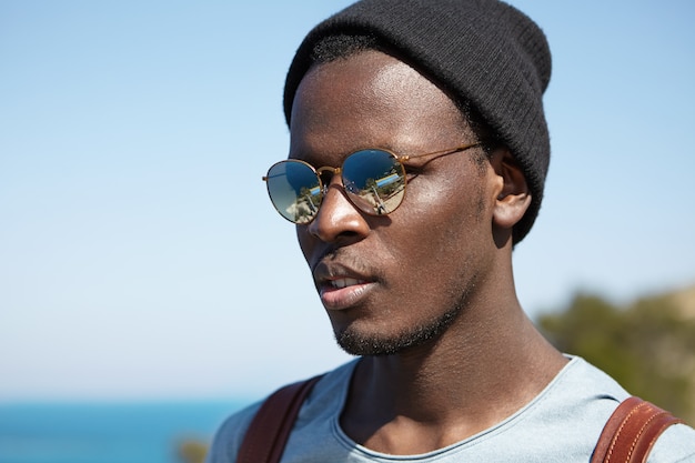 Handsome black young hipster wearing stylish hat and mirrored lens round sunglasses admiring beautiful and happy moments of his journey in foreign country while traveling alone around the world