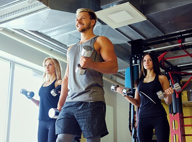잘생긴 금발의 운동하는 남성과 두 명의 날씬한 여성 피트니스 모델이 체육관 클럽에서 아령으로 어깨 운동을 하고 있습니다.