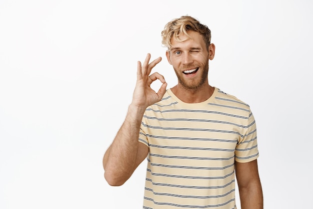 Free photo handsome blond bearded man with mascular body showing okay sign and winking smiling pleased standing over white background