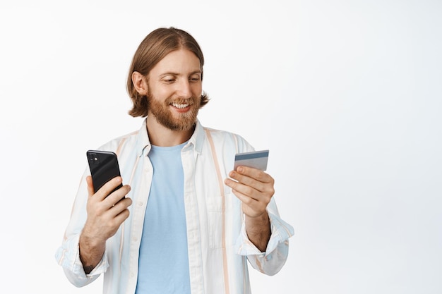 Handsome blond man with beard, paying online, purchase in internet with credit card and mobile phone, sending money with application, standing happy against white background