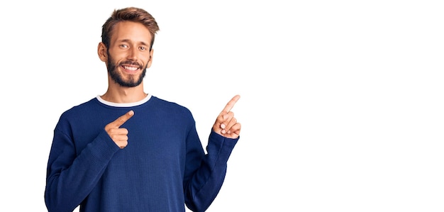 Handsome blond man with beard wearing casual sweater smiling and looking at the camera pointing with two hands and fingers to the side.