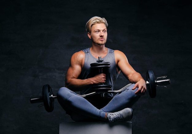 The handsome blond sporty male sits on a white box with crossed legs and holds dumbbell set.