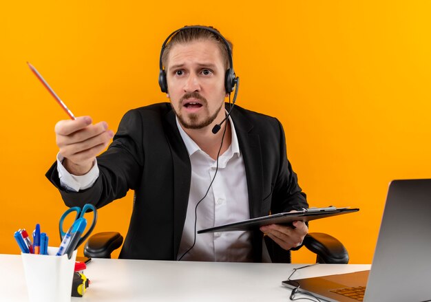 Handsome business man in suit and headphones with a microphone holding clipboard looking aside cofused and displeased sitting at the table in offise over orange background