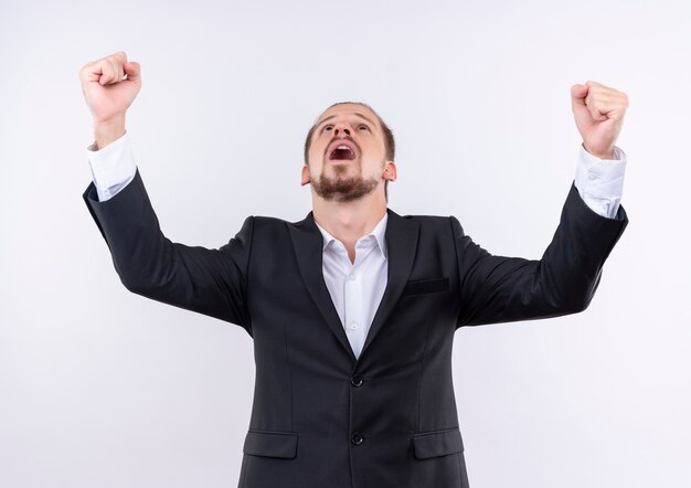 Handsome business man wearing suit clenching fists happy and crazy standing over white background