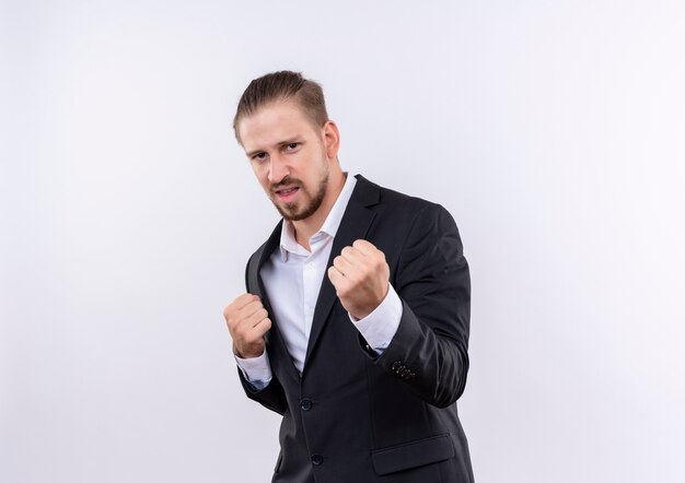 Handsome business man wearing suit clenching fists looking at camera with aggressive expression standing over white background