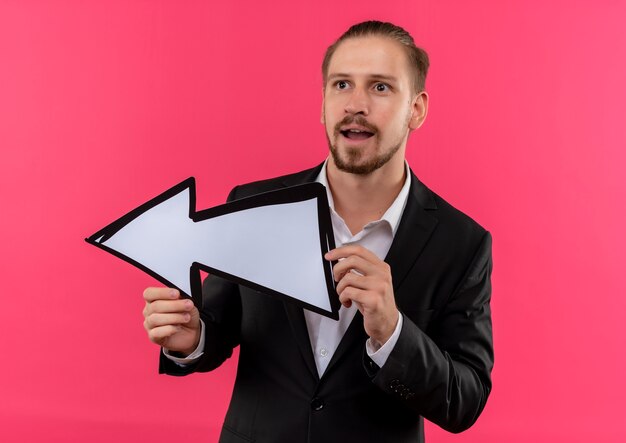 Handsome business man wearing suit holding arrow sign looking asie surprised standing over pink background