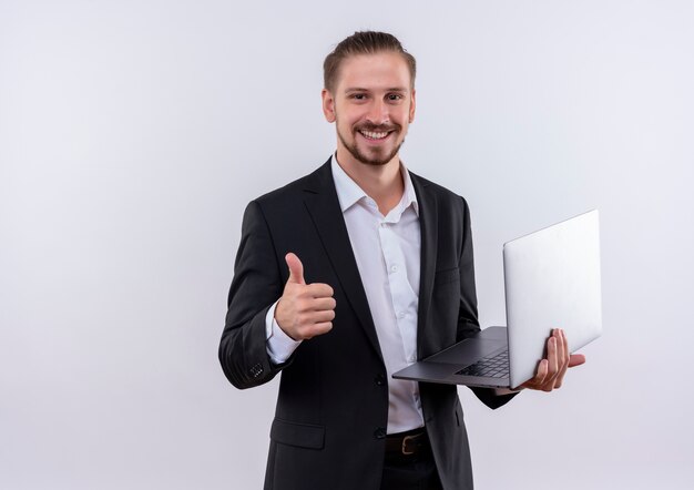 Handsome business man wearing suit holding blank pages looking at camera smiling cheerfully showing thumbs up standing over white background