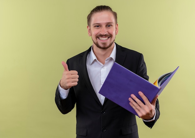 Handsome business man wearing suit holding blue folder looking at camera with smile showing thumbs up standing over green background